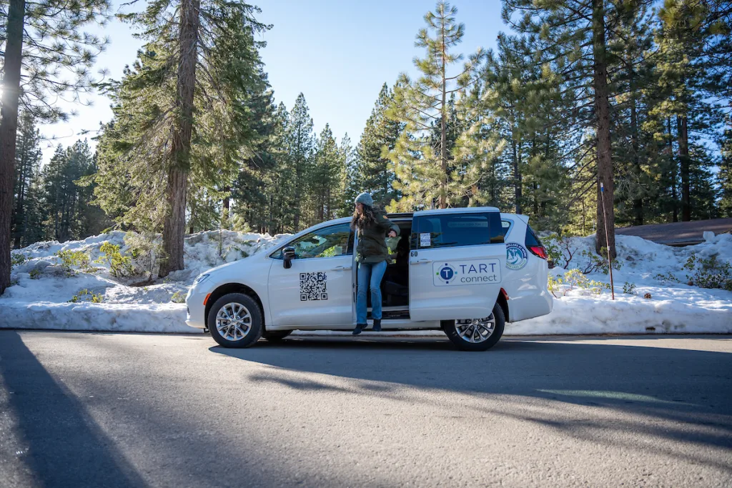 Person stepping out of a white van labeled 'TART connect' with a QR code, parked on a snowy road surrounded by pine trees under a clear blue sky.