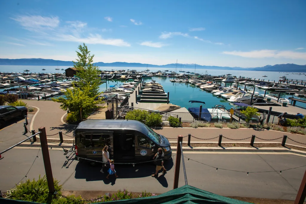 Black van parked near a marina with numerous boats docked in the water, surrounded by a scenic view of mountains and a clear blue sky, with two people walking nearby.